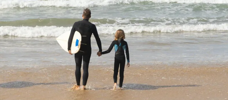 Dad and child wearing wetsuits walking into the sea with a surfboard 